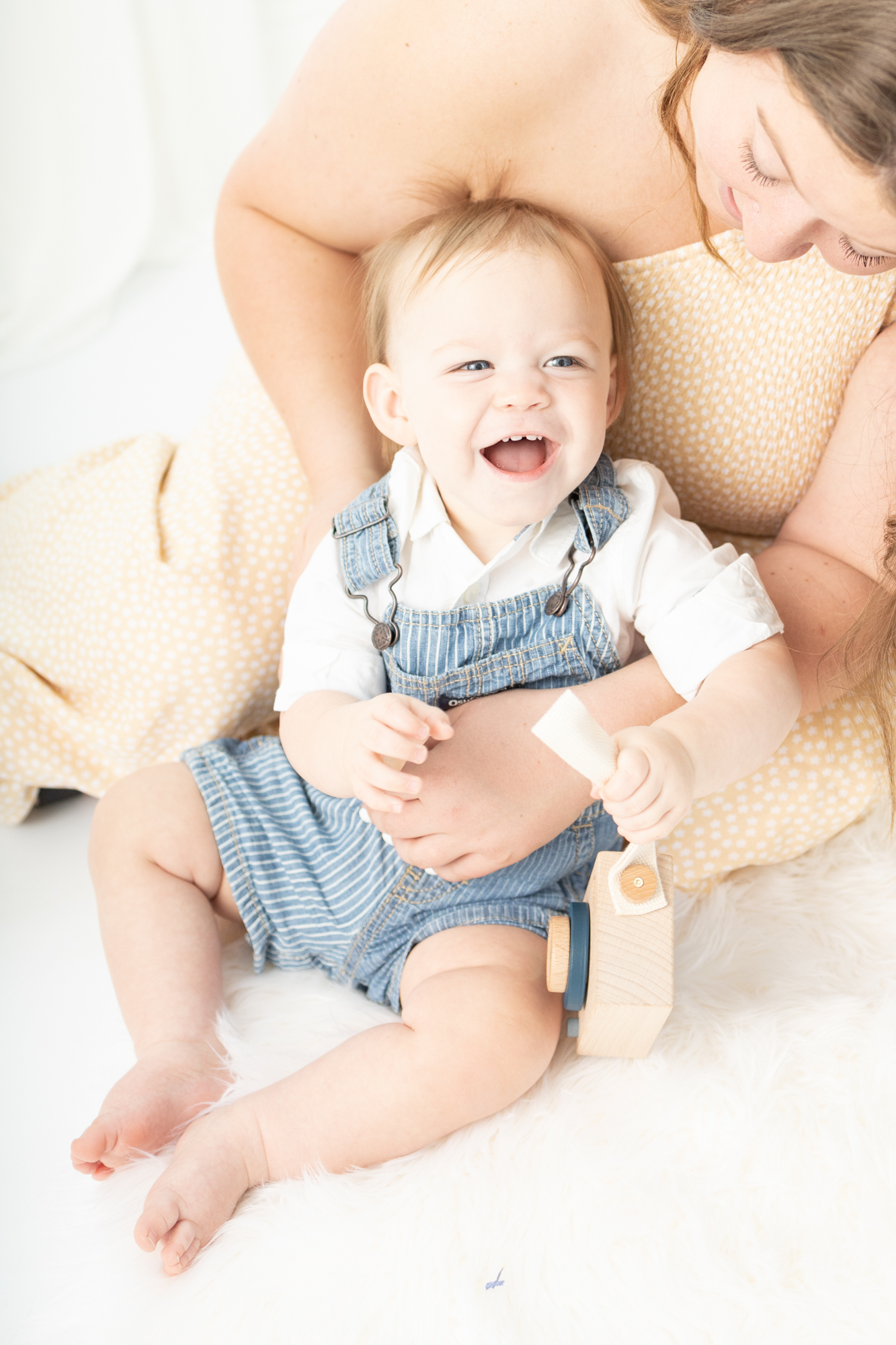 A laughing baby in overalls is tickled by mom on a bed in a studio