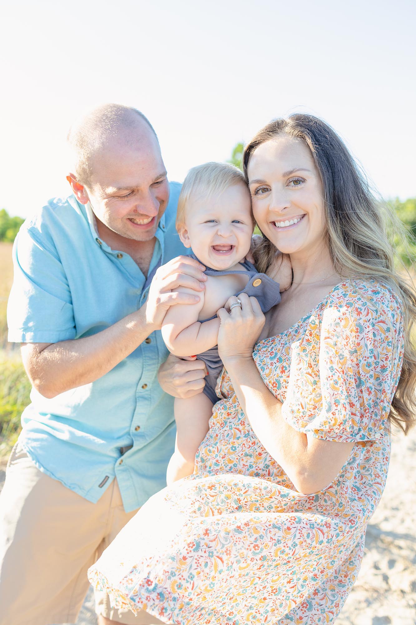 A giggling baby in overalls is tickled by mom and dad on a beach by Mary Eleanor Photography