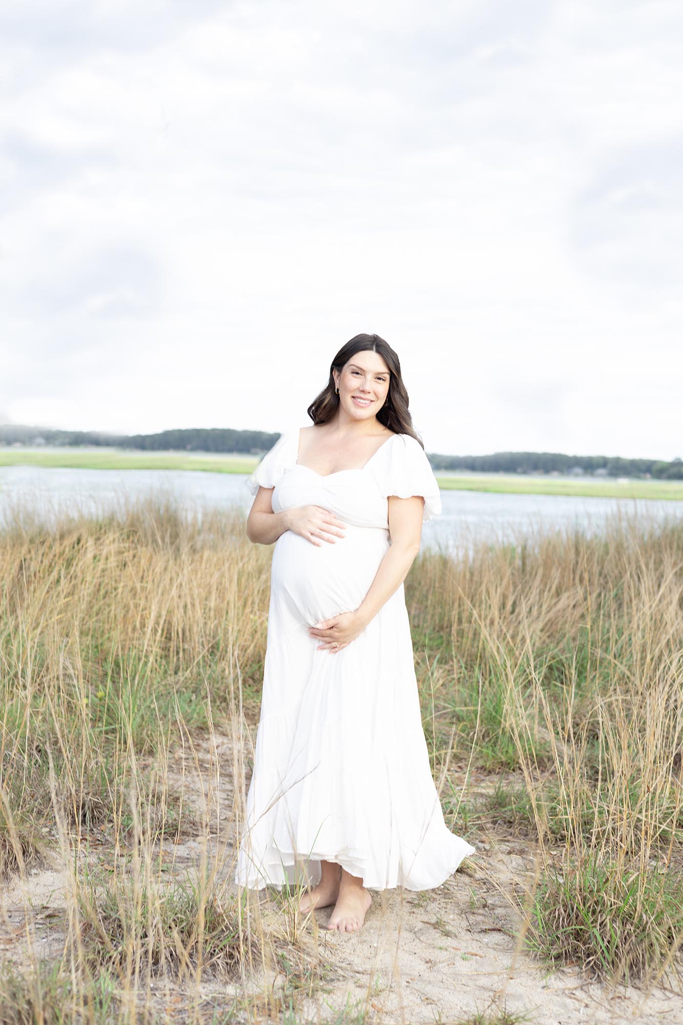 A smiling mom to be in a white maternity dress holds her bump on a beach after some Prenatal Yoga in Virginia Beach