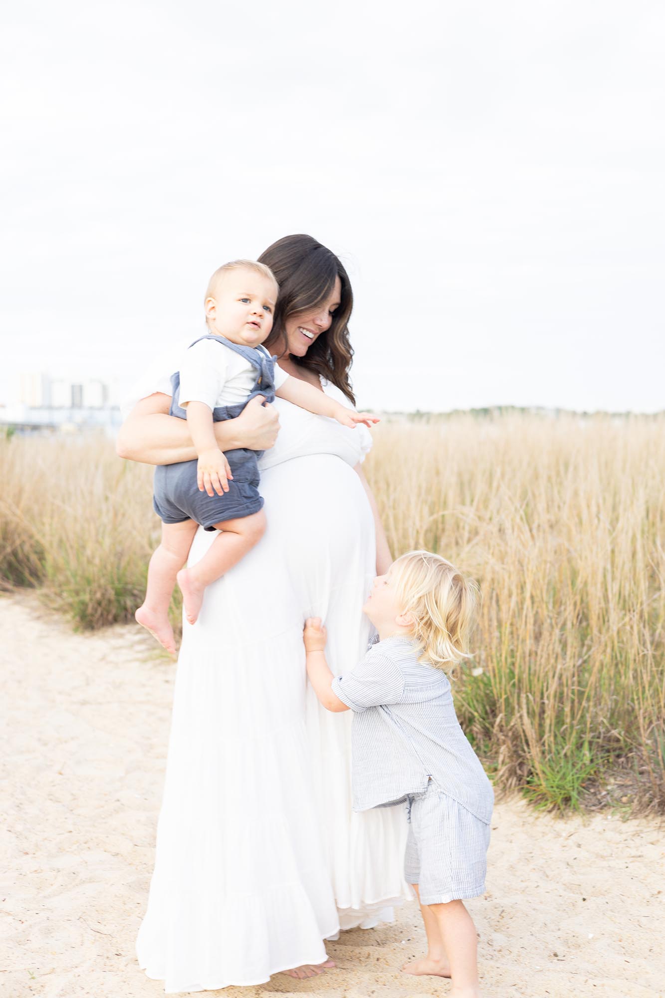 A smiling pregnant mom in a white maternity dress is hugged by her toddler while holding another on a beach after some Prenatal Yoga in Virginia Beach