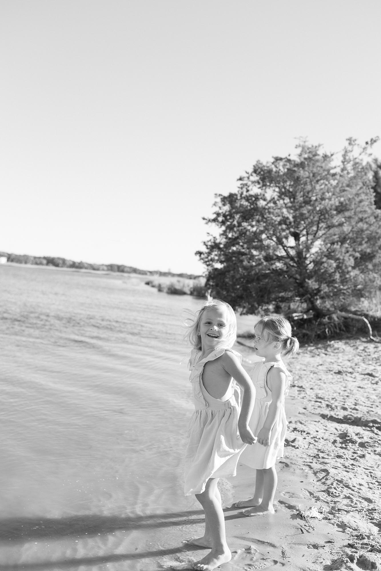 Happy toddler sisters in matching dresses play on the water's edge in black and white