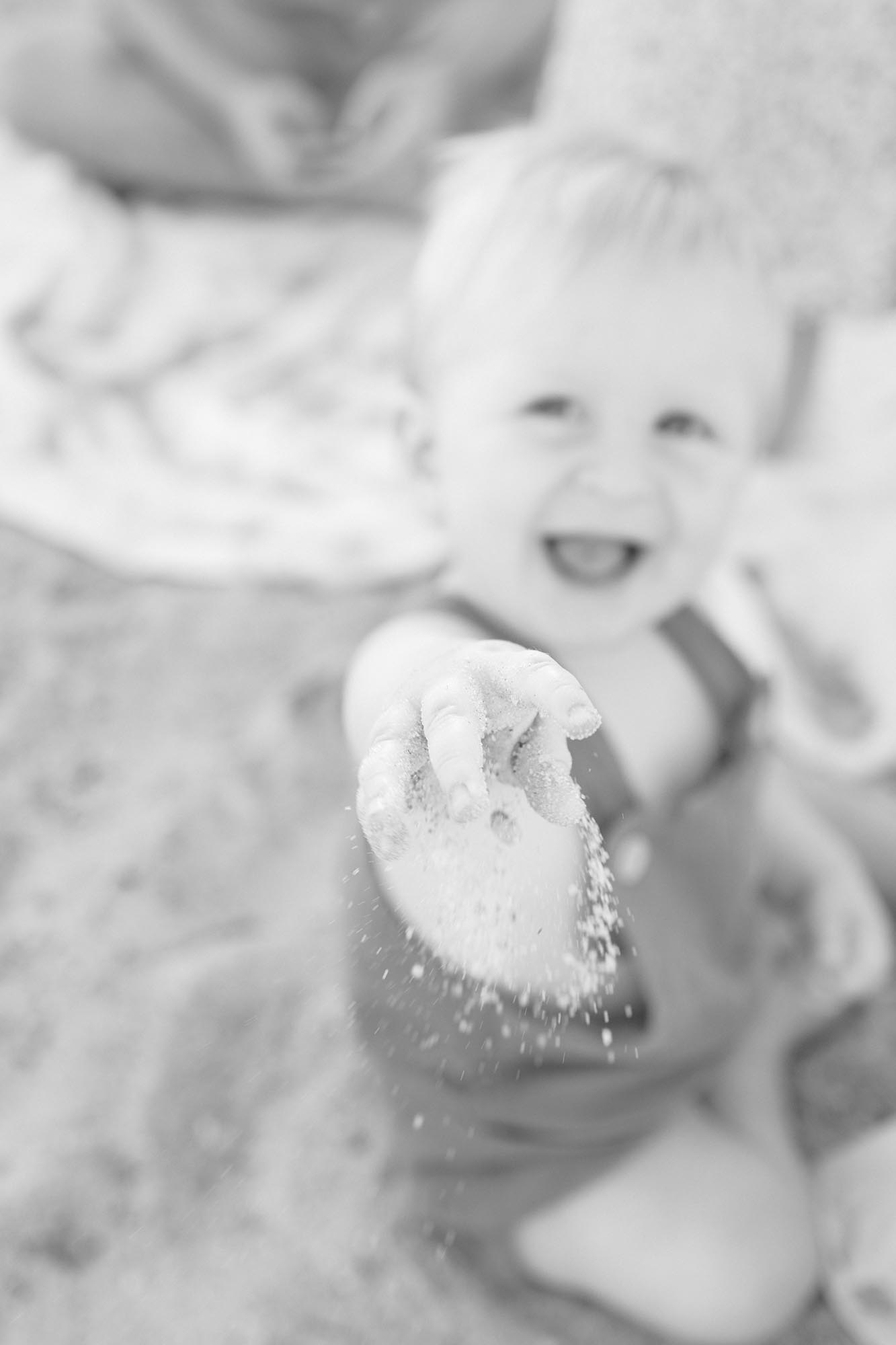 A happy toddler in overalls tosses sand in the air on a beach before visiting Summer Camps in Virginia Beach