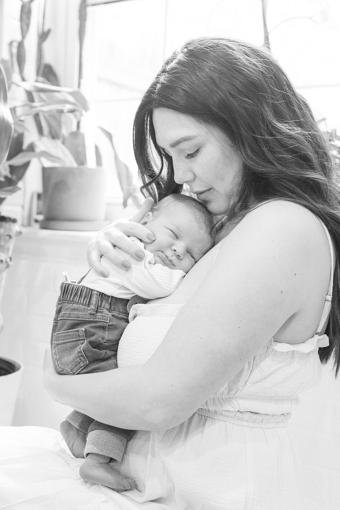 A new mom cradles her sleeping newborn against her chest while sitting under a window