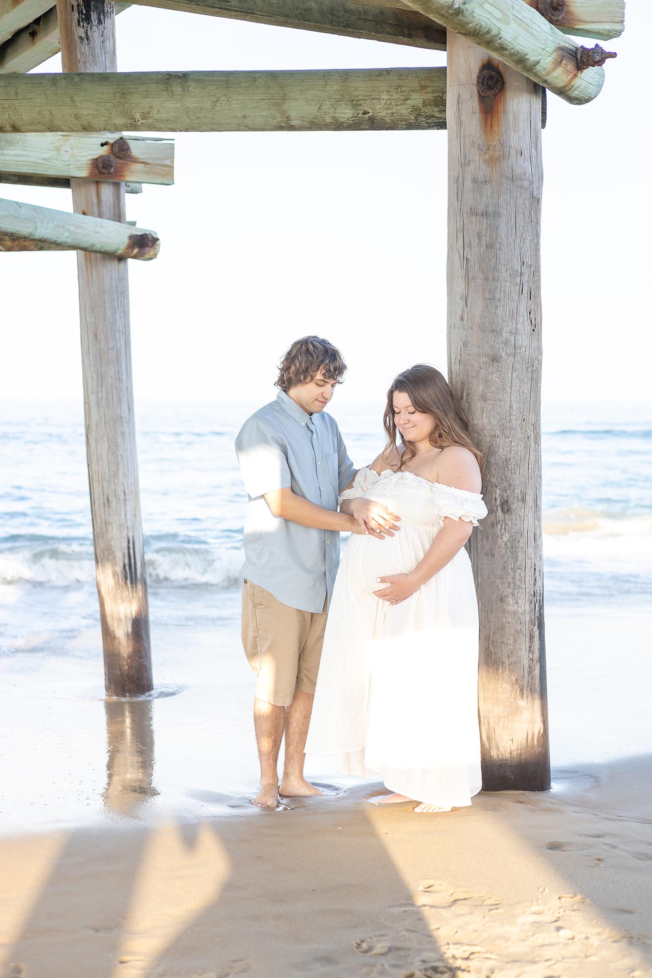 A mom to be in a white maternity gown leans on a pier piling on the beach with hands on her bump with dad