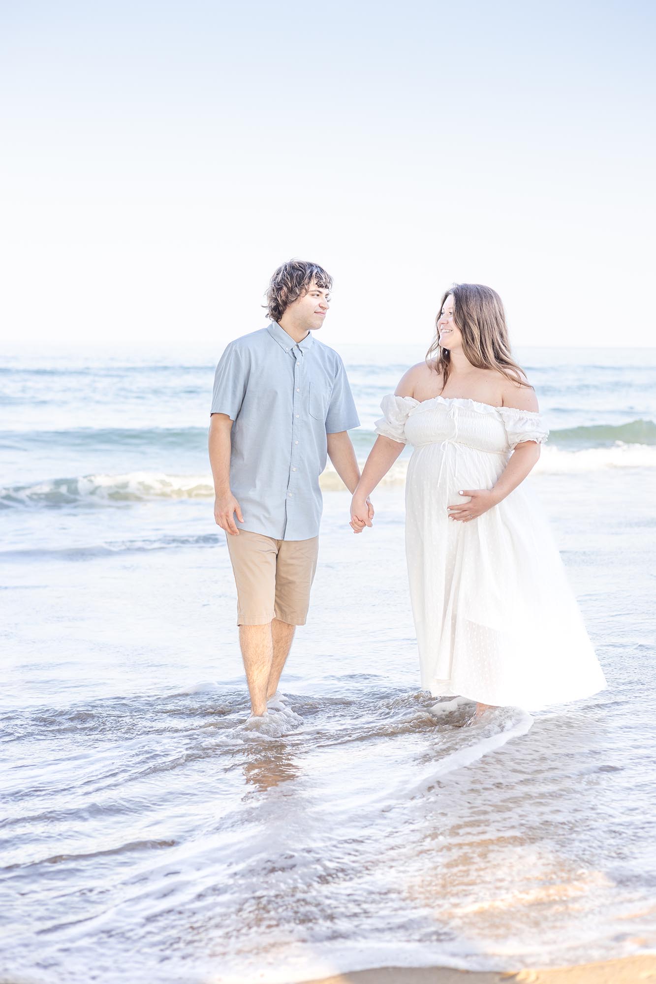 A pregnant couple wearing white and blue walk in the surf smiling at each other during one of the date night ideas in virginia beach
