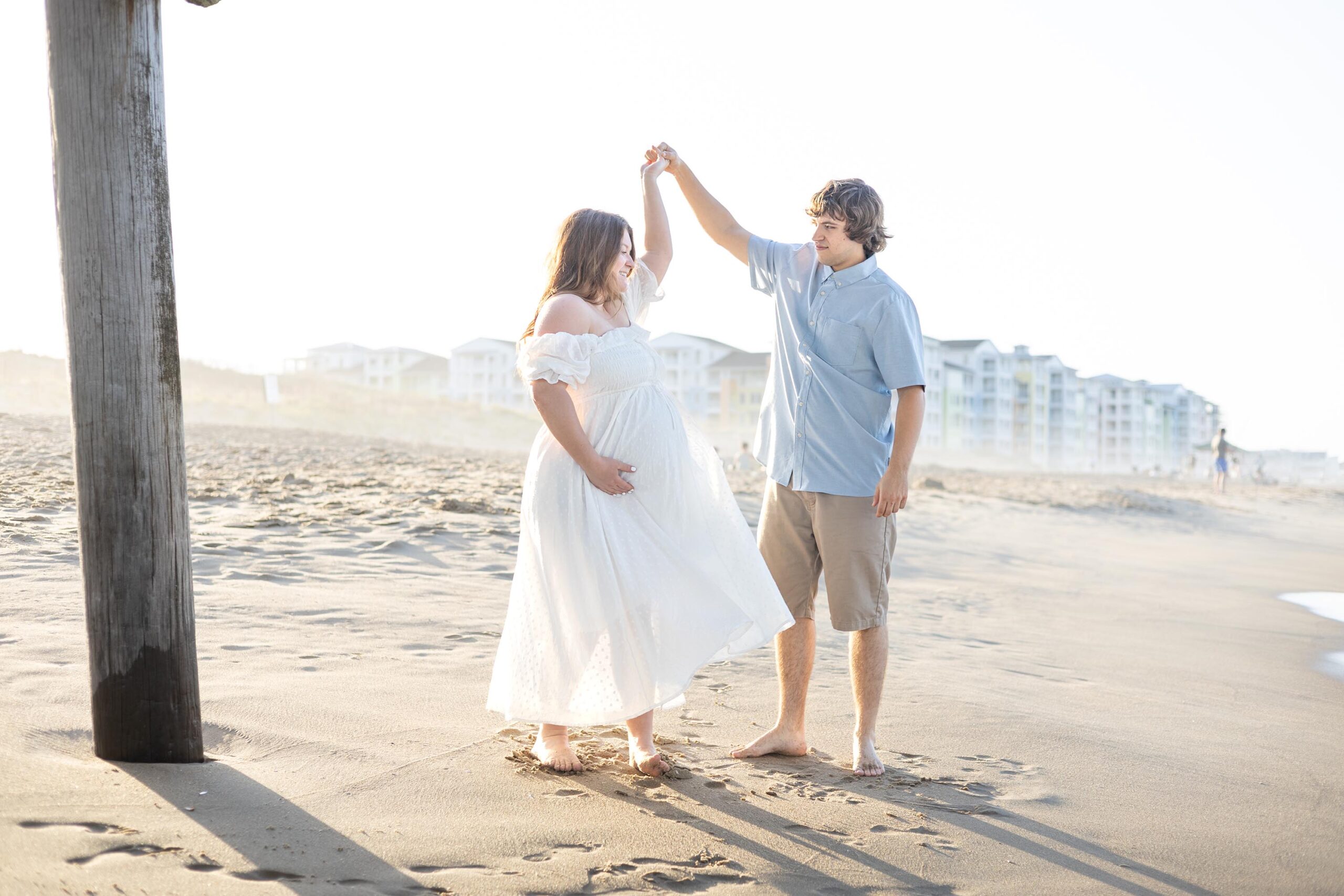 A happy expecting couple dances in white and blue on a beach under the pier at sunset during date night ideas in virginia beach