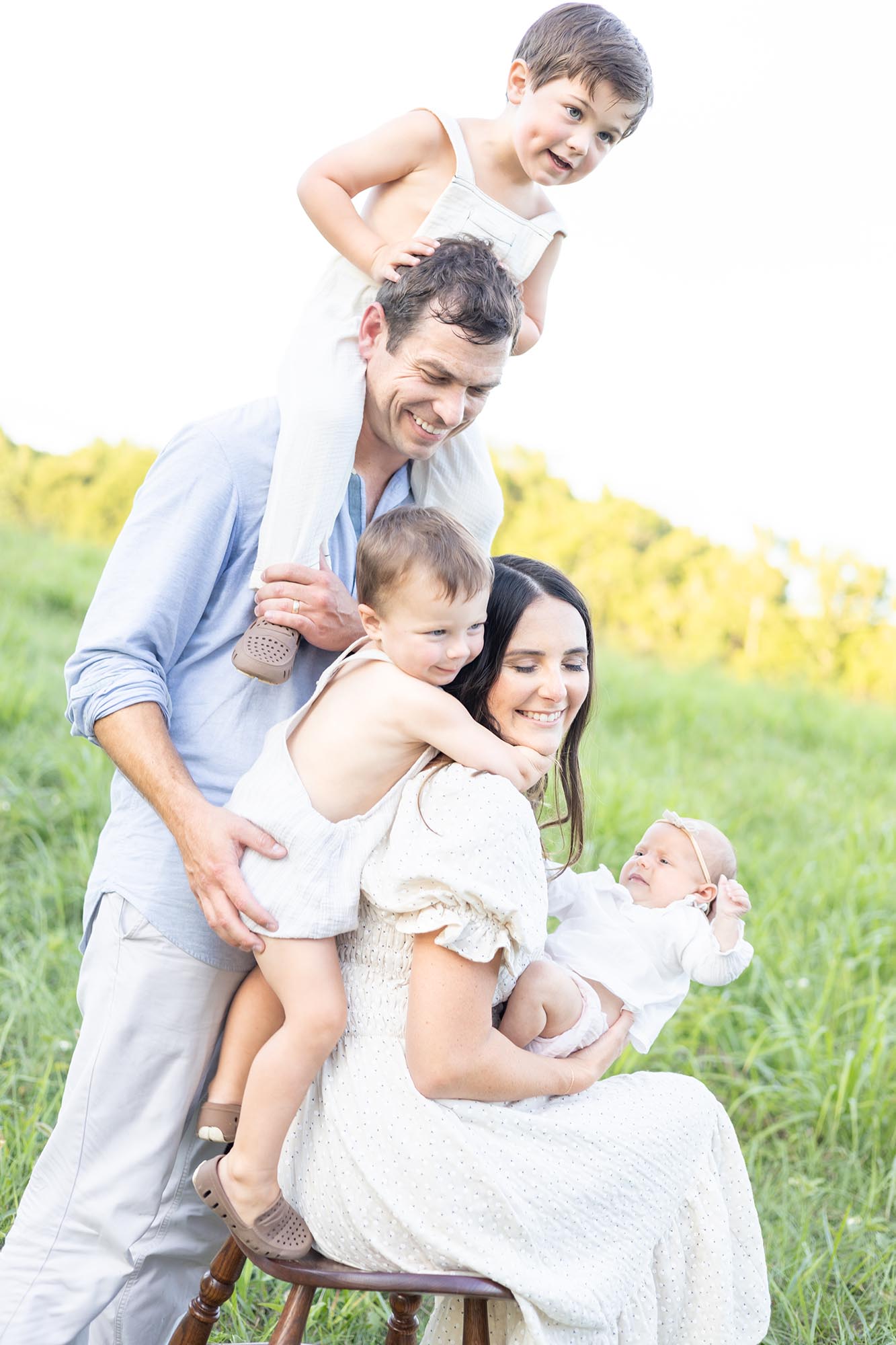 Two toddler brothers climb all over mom and dad while in a park field as mom holds baby sister before visiting farmers markets in virginia beach