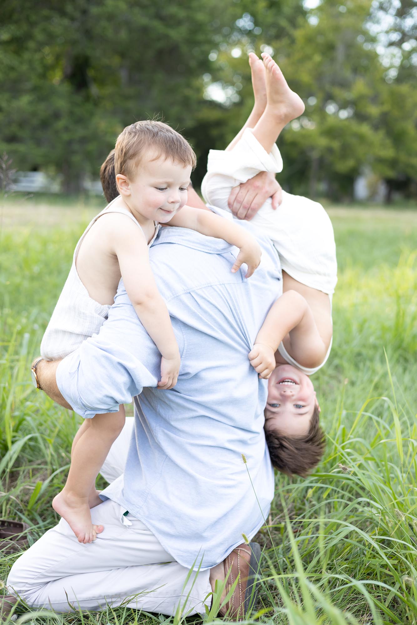 Toddler brothers in white overalls climb all over dad in a park field before visiting farmers markets in virginia beach