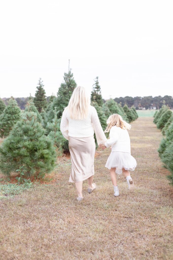A little girl holds her mother's hand and runs excitedly through the rows of Christmas trees.