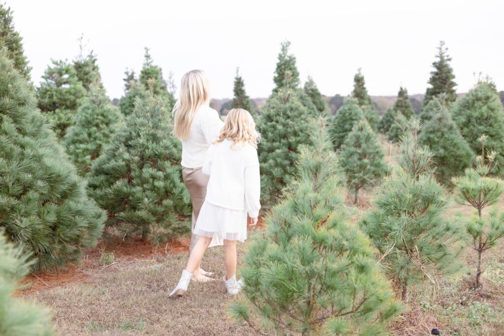 A Mother and Daughter wearing Cream knits and neutral skirts walk through a row of Christmas Trees holding hands during a Christmas Mini Sessions in Virginia Beach