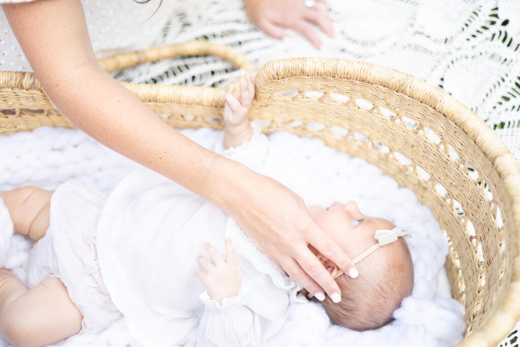 Infant in a white ruffled gown rests in a moses basket while mom brushes her cheek during Fall Activities in Virginia Beach. 