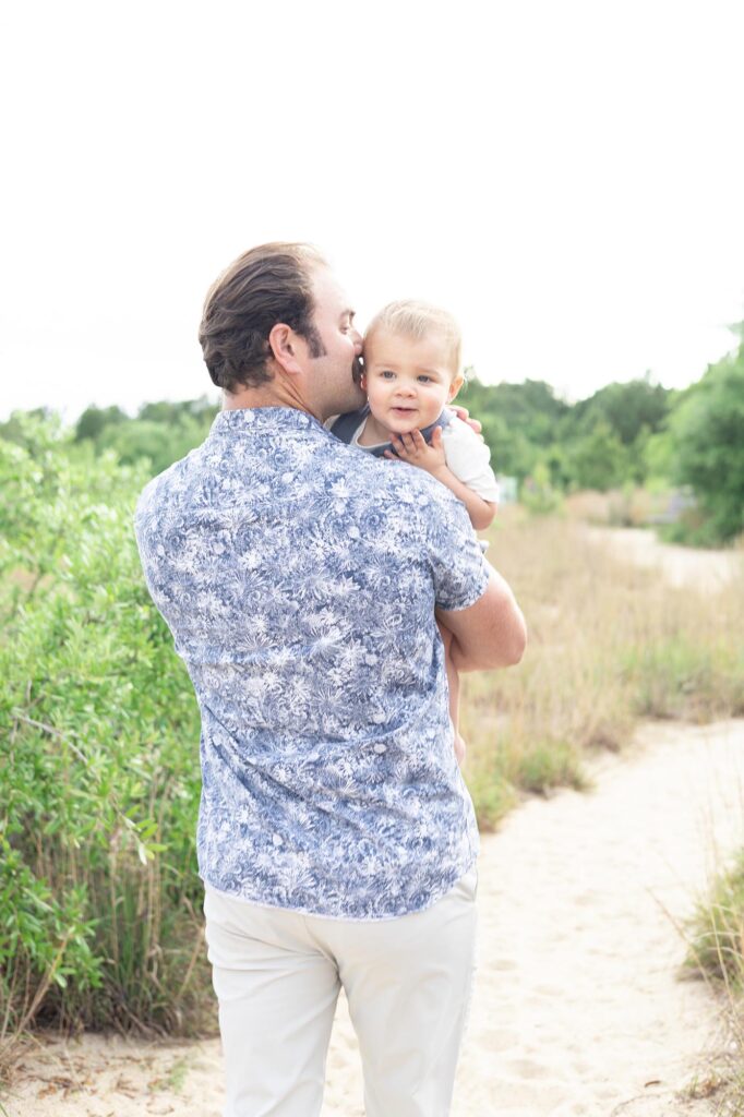 A father kisses his one year old son while walking down a beach path during a family vacation in Virginia Beach by Mary Eleanor Photography.