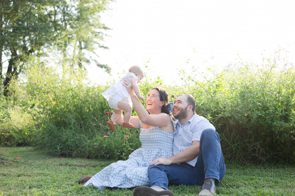 Mom in a blue gingham Nap Dress and Dad Sit together on the ground smiling while mom lifts baby in a white dress in the air after enjoying 4 Spots for Mini Golf in Virginia Beach.