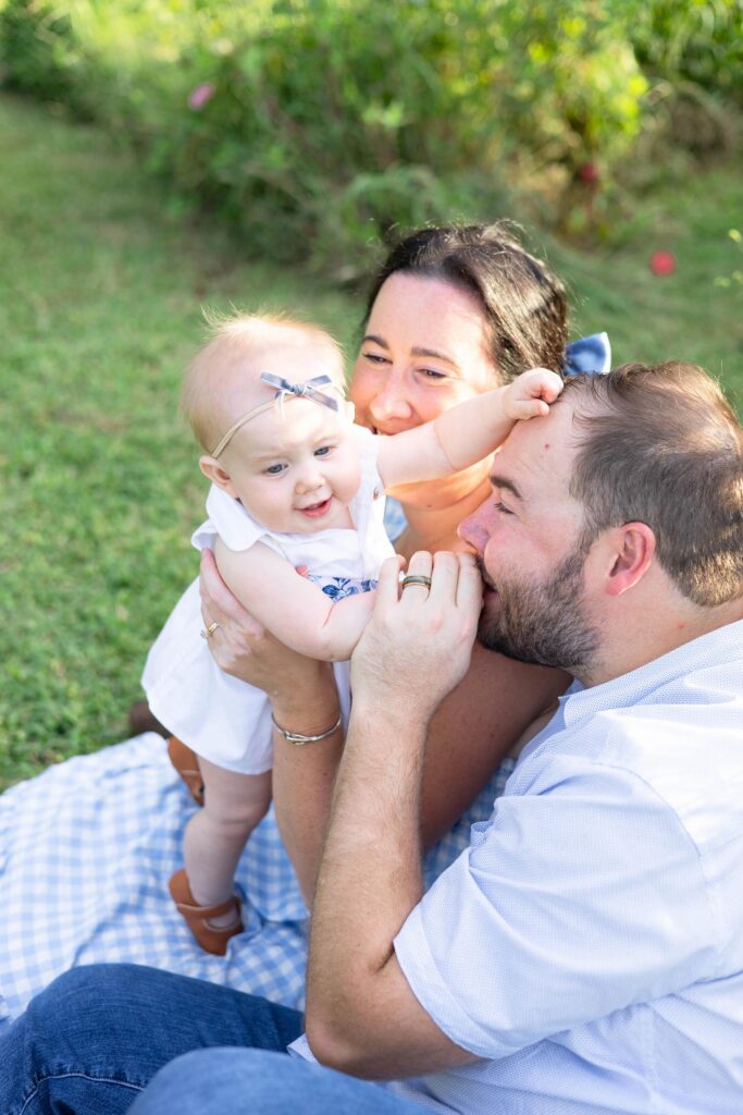 Mom and Dad Playing with their baby daughter while she reaches for dad and he kisses her hand after enjoying 4 spots for mini golf in Virginia Beach.