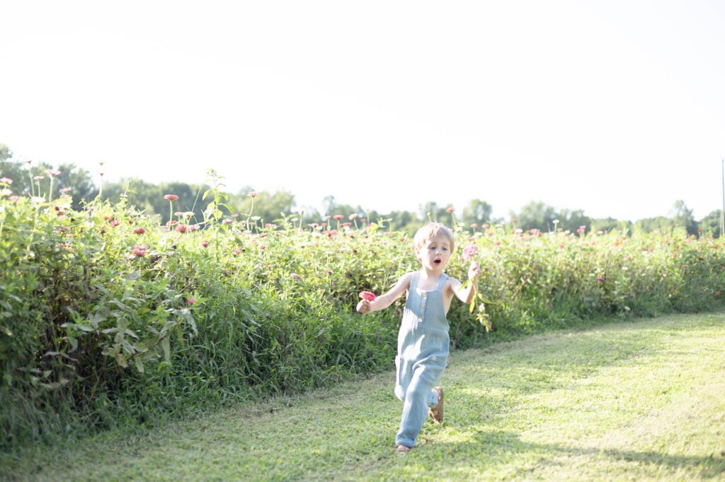 Little boy wearing blue overalls runs through a field holding flowers in each hand at a Playgrounds in Virginia Beach during a Family session by Mary Eleanor Photography.
