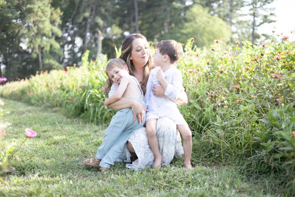 A mom wearing a white dress with small blue flowers snuggles her two little boys on her lap by Mary Eleanor Photography.