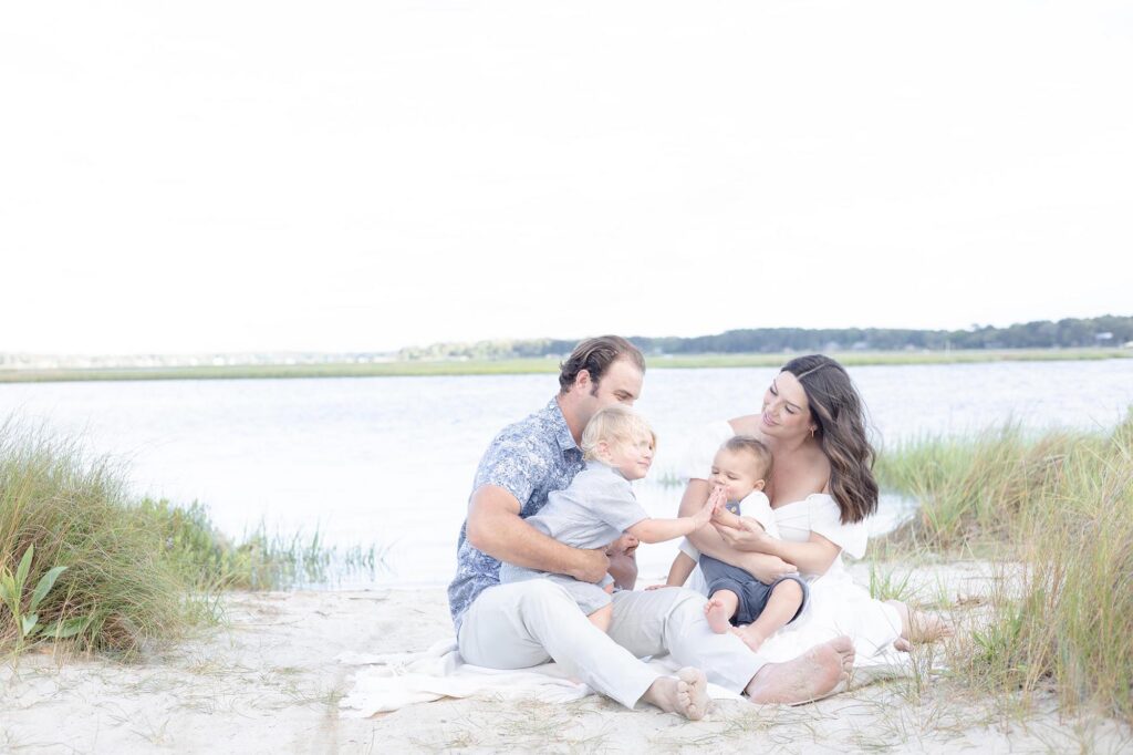 A Mom in a white off the shoulder dress and Dad in a blue floral pattern short sleeve button down sit on the beach holding their toddler and baby playing together during Family Photos Virginia Beach by Mary Eleanor Photography.
