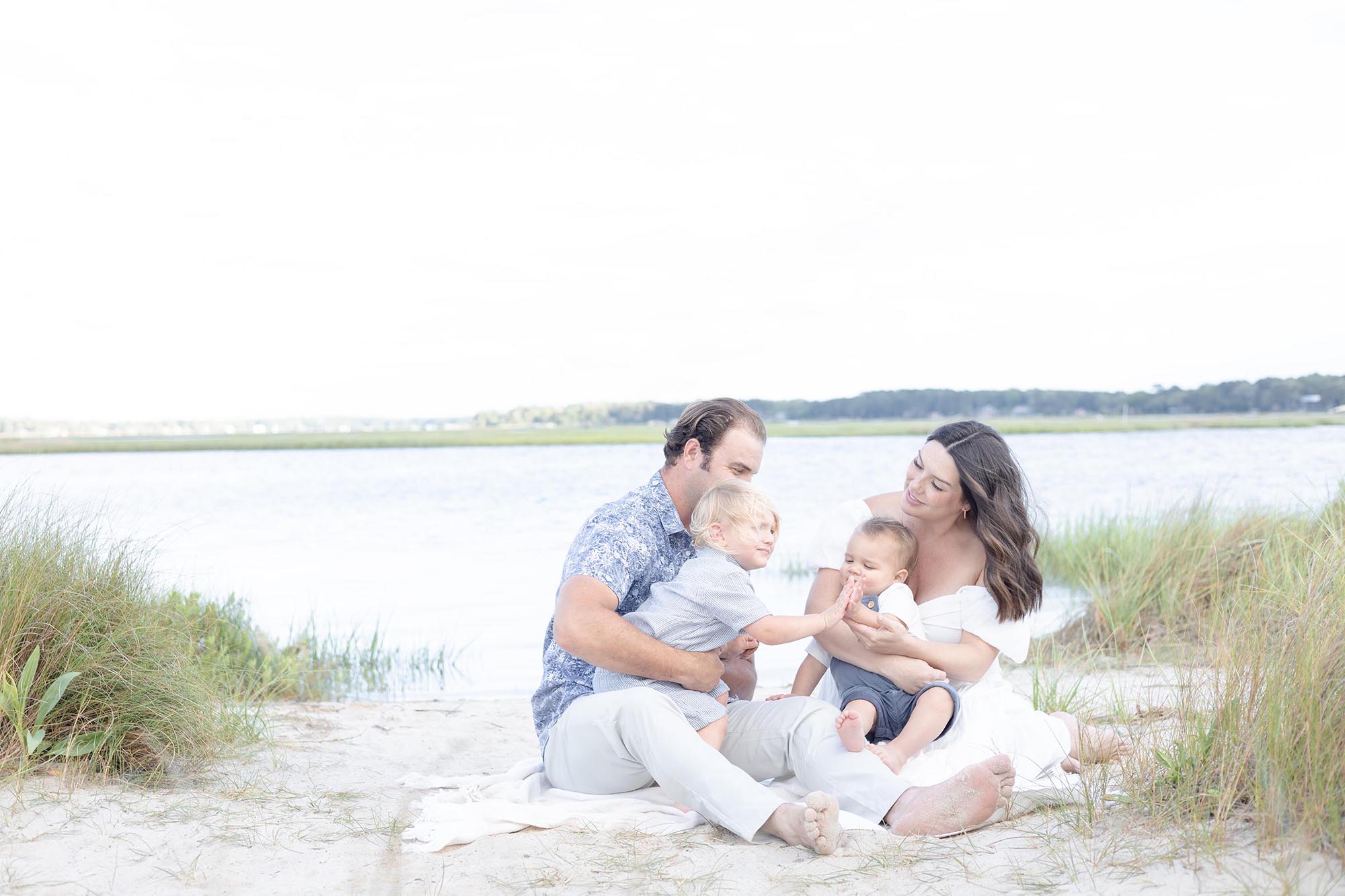 A Mom in a white off the shoulder dress and Dad in a blue floral pattern short sleeve button down sit on the beach holding their toddler and baby playing together during Family Photos Virginia Beach by Mary Eleanor Photography.
