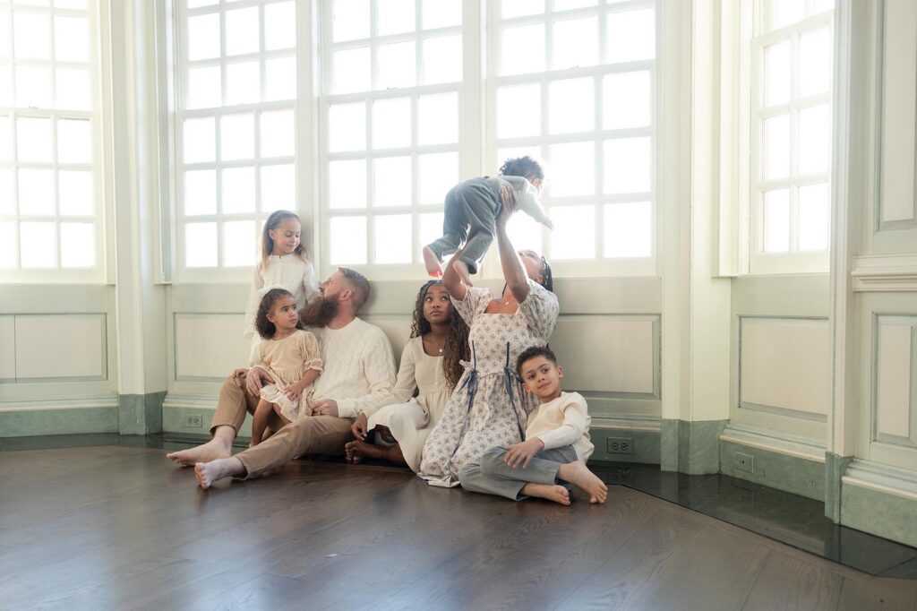 A family of 7 sits together on the floor, playing while mom tosses the littlest in the air during a family session after visiting Children's Museum Portsmouth.