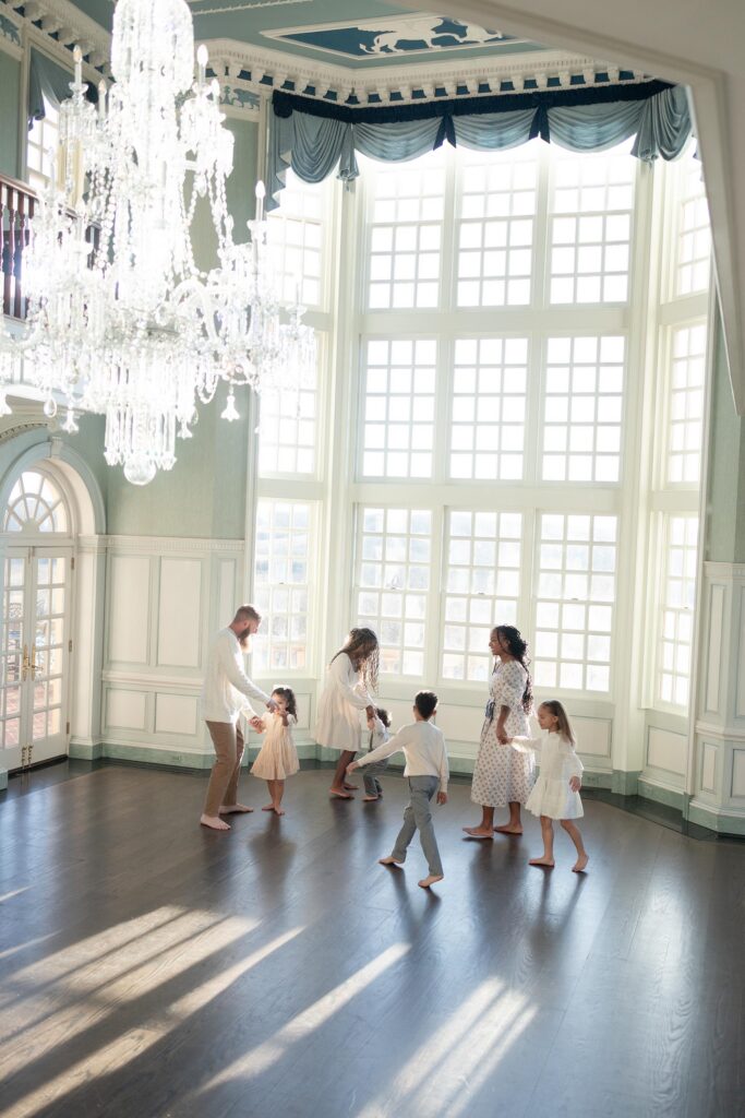 A family with Mom, Dad and 5 children has a dance party together in a large room with floor to ceiling windows by Mary Eleanor Photography.