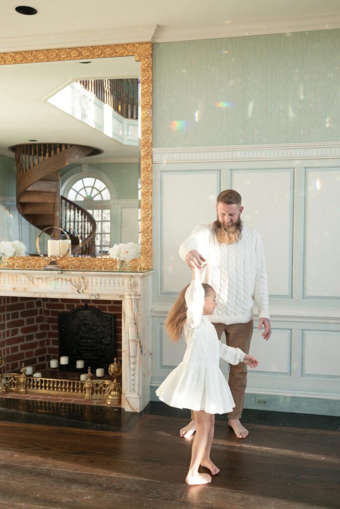 A Dad in a cream cable knit sweater twirls his young daughter during a family session by Mary Eleanor Photography.