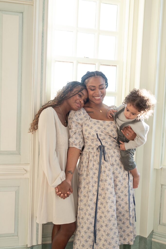 A Mom in a white dress with small blue flowers holds hands with her oldest child while holding her youngest on her hip after Children's Museum Portsmouth.