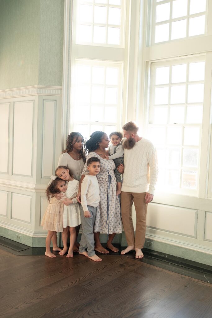 A family of 7 snuggles close together next to a large window by Mary Eleanor Photography.