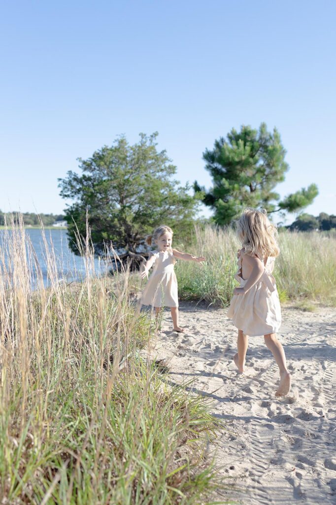 Two little girls in matching cream smock dresses run toward each other on the beach during Family Photos Virginia Beach by Mary Eleanor Photography