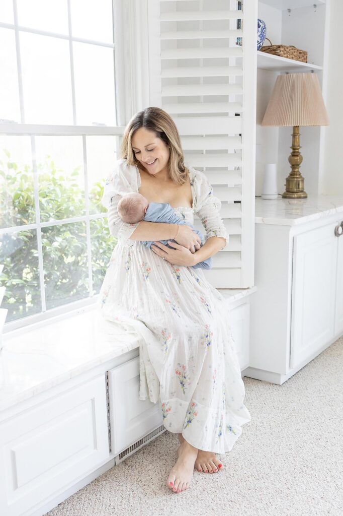 A new Mom cradles her newborn baby while sitting on a window seat wearing the DÔEN painted blooms Bijou dress. One of my favorite dress brands for Moms during an in-home session by Virginia Beach Newborn photographer Mary Eleanor Photography.