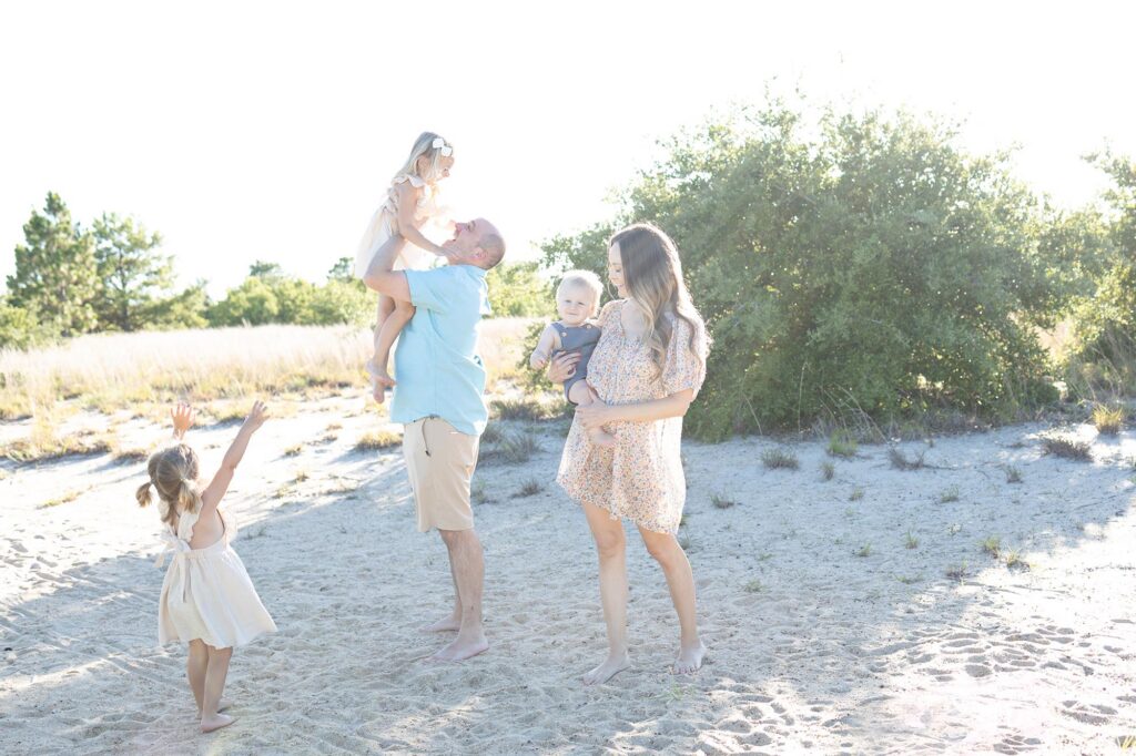 A mom in a floral dress holds her one year old and looks at her young daughter while the dad tosses their older daughter up in the air before Virginia Beach Fireworks by Mary Eleanor Photography.