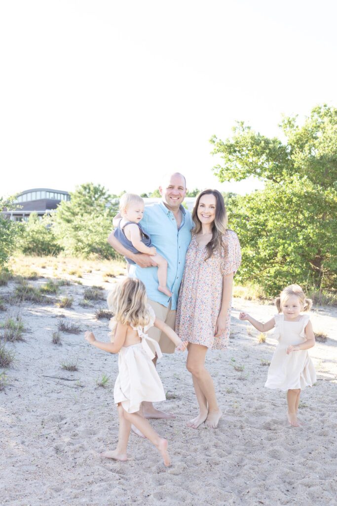 A Mom and Dad stand together in the sand with Dad holding their one year old son while their young girls run around them playing during a Family session by Mary Eleanor Photography before Virginia Beach Fireworks.