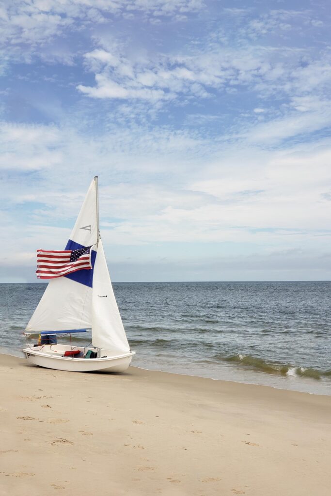 A small white sailboats sits on the sand at the beach with its blue and white sail up and an American Flag waving from the mast before Virginia Beach Fireworks.
