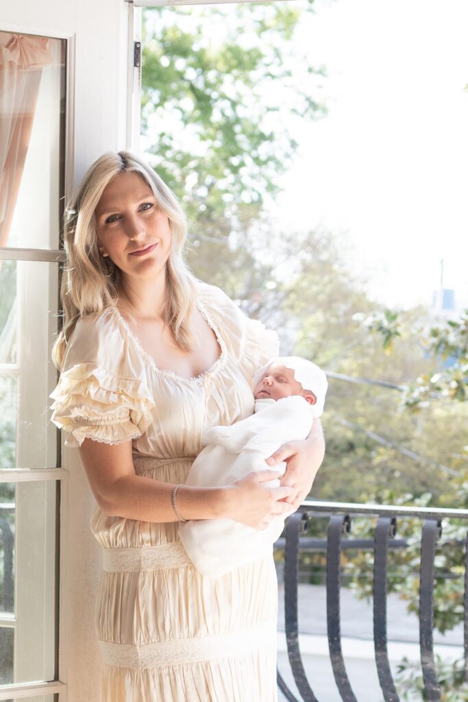 A Mom stands at the door of a balcony cradling her newborn while wearing a champagne colored satin and lace dress by Christy Dawn one of My Favorite Dress Brands for Mom during In-home Virginia Beach Newborn Photos by Mary Eleanor Photography.
