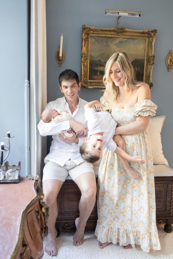A Mom and Dad hold their children and play with them during an in-home newborn session by Virginia Beach Newborn photographer Mary Eleanor Photography. Mom wears a blue and peach floral pattern dress by Nothing Fits But One of My Favorite Dress Brands for Moms.