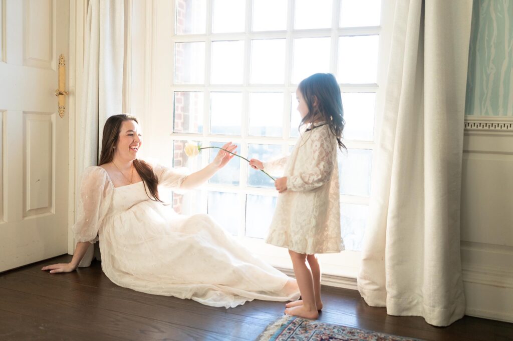 Mom sits on the floor next to a window wearing a dress by Christy Dawn one of my favorite dress brands for mom while her little girl playfully hands her a rose.