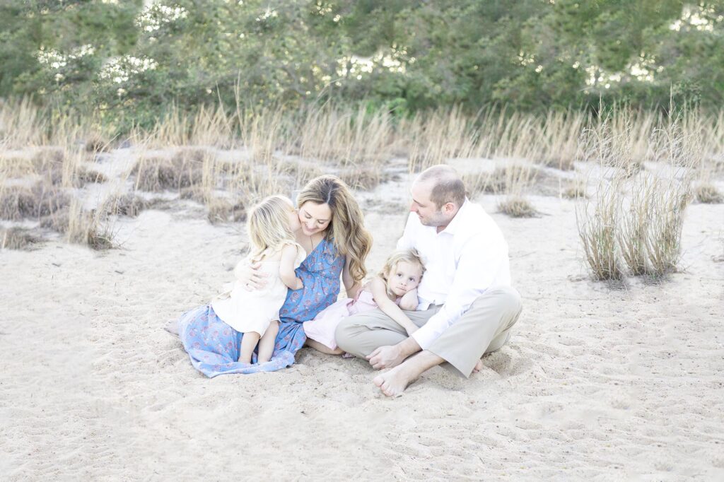 A family of four, Mom, Dad, and Two little girls, sits together on the sand in front of coastal beach grasses during Virginia Beach Maternity Photos by Mary Eleanor Photography.