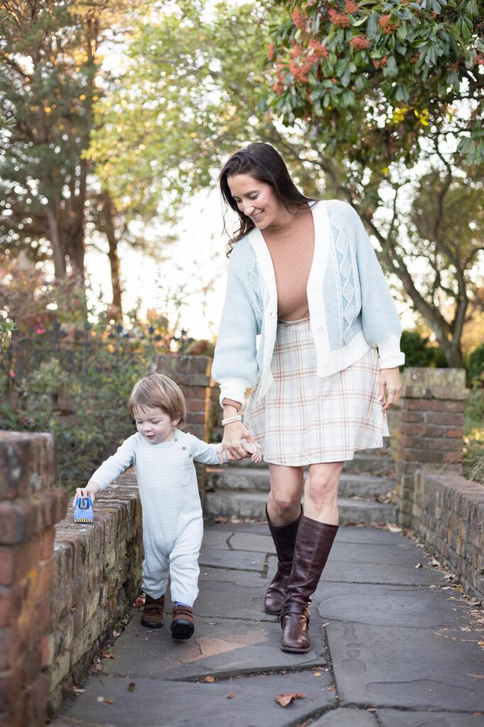 A Mom in a Tan Sweater and cream and white skirt with a pale blue and white cardigan walks along a path holding her son's hand while he drives a small toy truck along a brick wall during their Fall Family Photos Virginia Beach by Mary Eleanor Photography.
