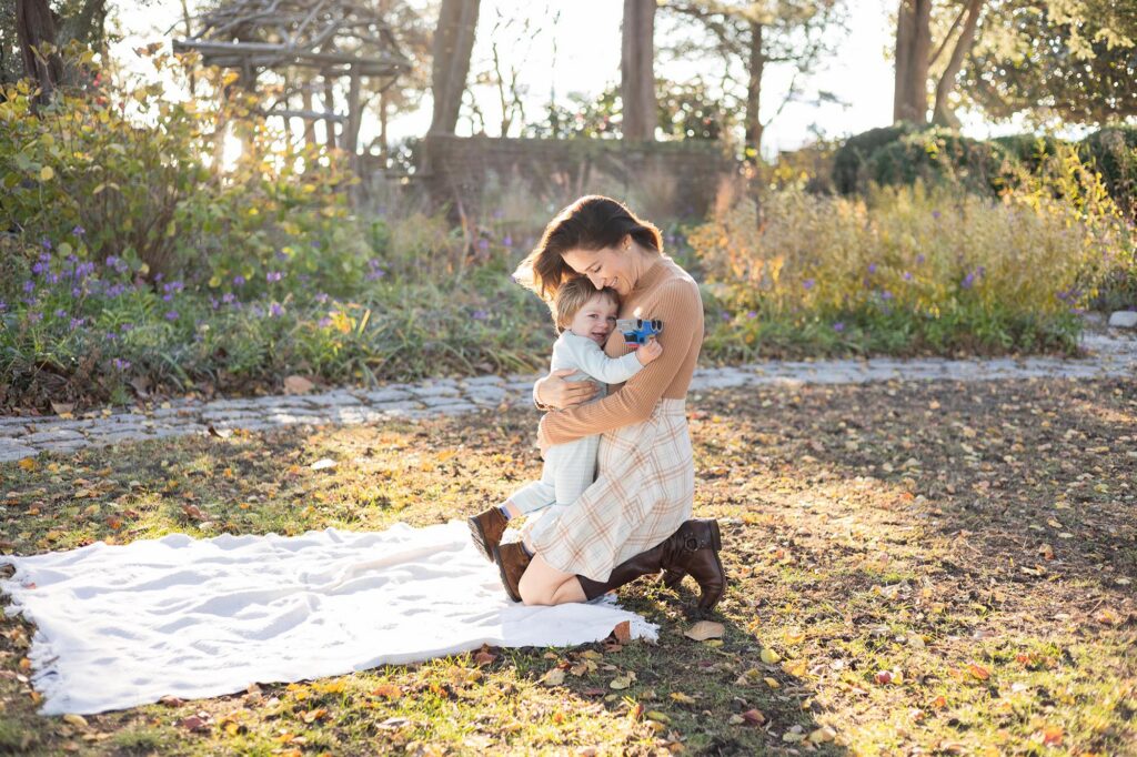 A Mom snuggles her son close smiling and laughing as they sit on a blanket in a fall garden during Fall Family Photos Virginia Beach by Mary Eleanor Photography.