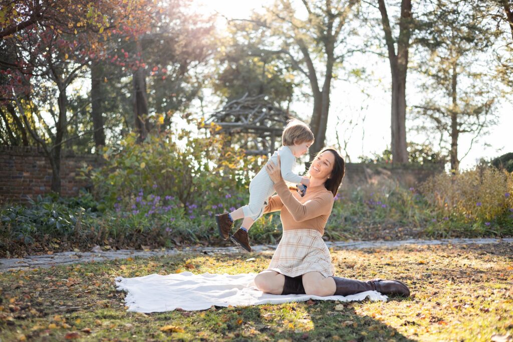 A Mother and son play, with Mom lifting him up in the air while sitting on a white blanket in a beautiful Fall Garden during fall Family Photos in Virginia Beach by Mary Eleanor Photography.