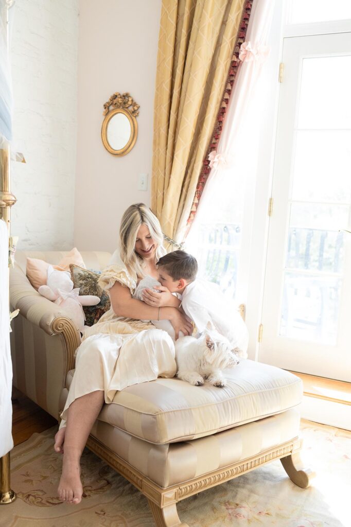 Mom Sits on a chaise while holding her newborn and her toddler kisses baby during newborn siblings with photos.